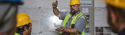 Instructor wearing a yellow hard hat and reflective safety vest gestures while explaining information on a whiteboard to a group of workers in a training setting