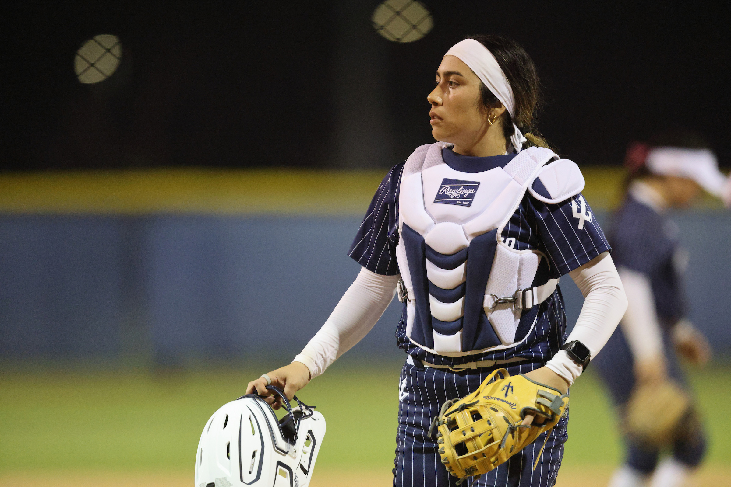 UC San Diego softball player Lily Hermosillo, wearing a Tritons uniform and catcher vest, holds a helmet while gazing into the distance.